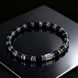 May include: A black beaded bracelet with silver-colored accents. The bracelet features cylindrical black beads separated by silver-toned rings. A silver-colored, textured cylindrical bead is centered on the bracelet. The bracelet is displayed on a dark, reflective surface.