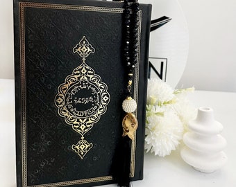 May include: A black leather-bound Quran with gold lettering and intricate designs. A black tassel with a gold charm hangs from the book. A black beaded prayer chain with a white pearl charm is attached to the book.