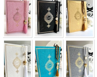 May include: Six different colored covers of a Quran with matching tassel and prayer beads. The covers are pink, black, gold, white, turquoise, and gray.