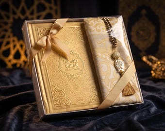 May include: A gold-colored Holy Quran book with intricate embossed patterns, packaged in a clear box with a matching gold ribbon. Beside the book is a folded cream-colored fabric with a gold tassel and a beaded prayer bead strand.