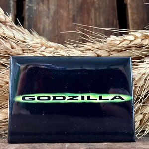Può includere: Bottone quadrato nero con un logo "GODZILLA" verde.