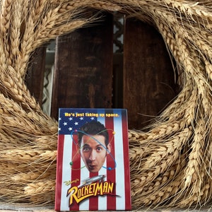 May include: A red, white and blue button featuring a man's face with the text "Disney's Rocketman" and the phrase "He's just taking up space."