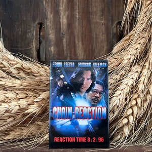 May include: A VHS tape case for the film "Chain Reaction" starring Keanu Reeves and Morgan Freeman. The case is black with a blue and white design. The text "Reaction Time 8:2:96" is printed on the bottom of the case.