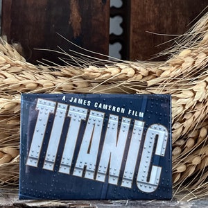 Può includere: Un magnete blu e argento con il testo "A James Cameron Film TITANIC" su di esso. Il magnete è su una superficie di legno con spighe di grano dietro.