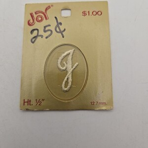 May include: A white embroidered letter J patch centered on a gold-colored rectangular card. The card features the Joy logo, the text 25 cents, 1.00, and Ht. 1/2 inch. The letter is enclosed within an oval outline printed on the card surface.
