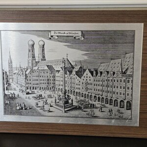 May include: A rectangular wall plaque featuring a silver-toned metal plate with a black and white historical etching of a city square. The image depicts the Marienplatz in Munich with the text Der Marckt zu München. It has a wood-grain frame.