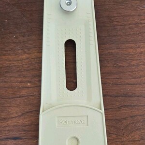 May include: A beige Kenmore sewing machine attachment featuring an elongated plastic body with a central slot, a metal adjustment knob, and a circular base with a black center hole. The text 3/8 is embossed near the base.