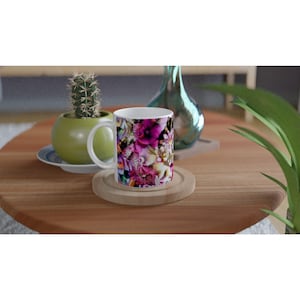 Pode incluir: Caneca de café de cerâmica branca com um design floral vibrante em tons de rosa, roxo e amarelo. A caneca está sobre um descanso de copo de madeira, com um cacto num vaso verde e um vaso decorativo no fundo.