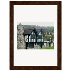 Könnte beinhalten: Gerahmter Druck mit einem historischen Gebäude mit Strohdach, schwarz-weißem Fachwerk und grünen Fensterläden. Das Bild ist in einem dunkelbraunen Holzrahmen gerahmt. Ein steinerner Schornstein und Grabsteine sind sichtbar.