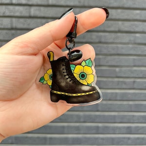 May include: Black boot-shaped keychain with yellow accents, featuring a floral design. The keychain has a black clasp and a yellow loop. The boot has yellow stitching and a yellow sole.