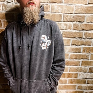 Puede incluir: Sudadera con capucha gris oscuro con bolsillo delantero y capucha con cord&oacute;n. La sudadera con capucha presenta un gr&aacute;fico de tres dados con el n&uacute;mero siete. Los dados est&aacute;n rodeados de peque&ntilde;as estrellas. La sudadera con capucha la lleva una persona.
