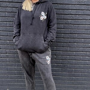 Puede incluir: Un conjunto a juego de sudadera con capucha y pantal&oacute;n de ch&aacute;ndal gris carb&oacute;n. La sudadera con capucha tiene una capucha con cord&oacute;n y un bolsillo delantero. Ambas piezas presentan un gr&aacute;fico de dados con el n&uacute;mero siete. La persona lleva zapatillas blancas y negras.