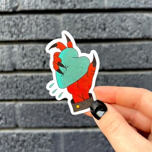 May include: A sticker featuring a red hand with black claws holding a teal heart. The hand has a black cuff with a gold button. Water droplet graphics are below the hand. The sticker is held by a person.