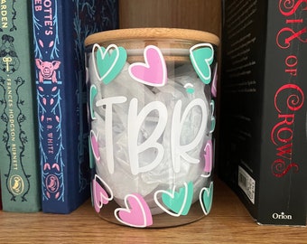 Könnte beinhalten: Ein klares Glasgefäß mit einem Bambusdeckel, verziert mit rosa und türkisfarbenen Herzen und dem Wort "TBR" in weißen Buchstaben. Das Gefäß steht auf einem Bücherregal mit anderen Büchern.