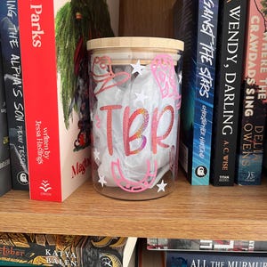 May include: A clear glass jar with a bamboo lid and a pink and white design that says "TBR" on the front. The jar is sitting on a bookshelf with other books.