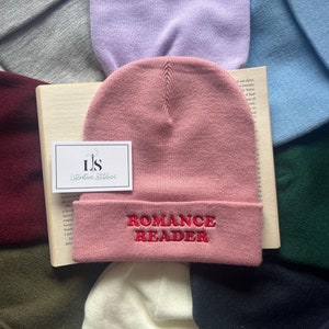 May include: A pink beanie with the words "ROMANCE READER" embroidered in red on the cuff. The beanie is surrounded by other beanies in various colours.