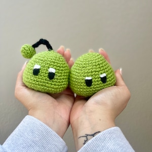 May include: Two green crocheted plushies with black eyes and white accents. One plushie has a black crocheted antenna.