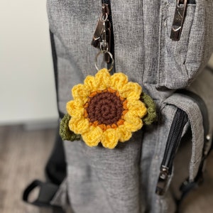 May include: A crocheted sunflower keychain with green leaves attached to a silver zipper pull on a gray backpack.