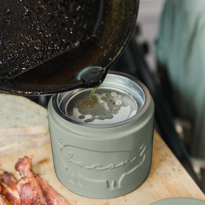 May include: A gray ceramic container with a pig design and the word "Bacon" embossed on the side.  The container is being filled with hot oil from a black cast iron skillet.