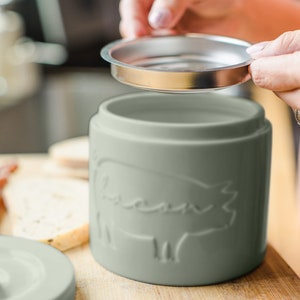 May include: A sage green ceramic canister with a pig design and a stainless steel lid. The canister is on a wooden surface.