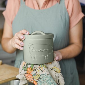 May include: A ceramic container with a lid, featuring a pig design and the word "bacon" embossed on the front. The container is a light green color.