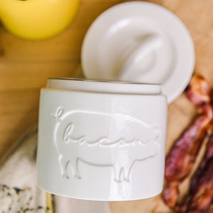 May include: White ceramic jar with a pig design and the word "bacon" embossed on the side. The jar has a lid with a small knob on top. The jar is sitting on a wooden surface with bacon strips in the background.
