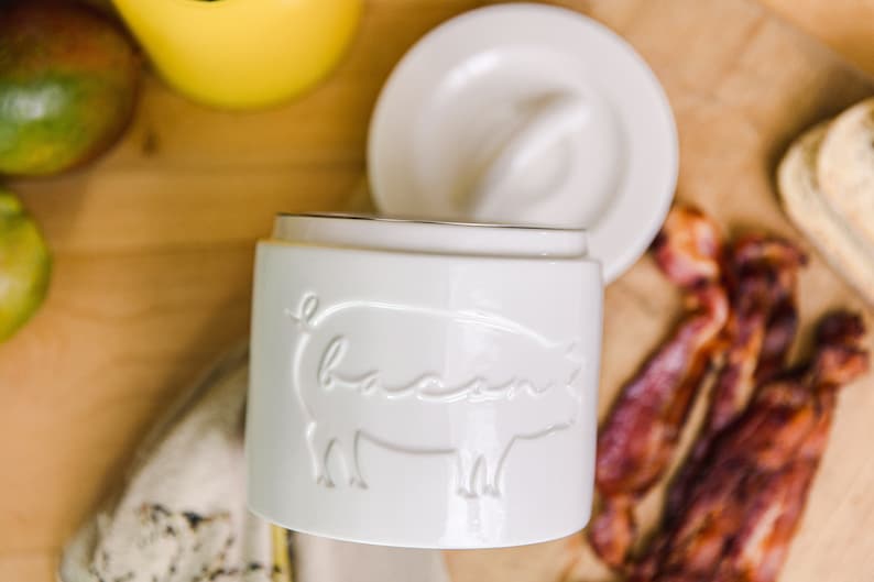 May include: White ceramic jar with a pig design and the word "bacon" embossed on the side. The jar has a lid with a small knob on top. The jar is sitting on a wooden surface with bacon strips in the background.