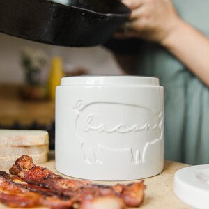 May include: White ceramic bacon jar with a pig silhouette and the word "bacon" embossed on the front. The jar is sitting on a wooden surface with cooked bacon strips beside it.