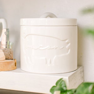 May include: White ceramic canister with a lid and a raised pig design. The canister is on a white shelf.