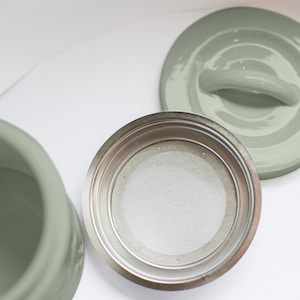 May include: A silver coffee filter with a mesh insert sits on a white surface. A sage green ceramic lid with a handle is also visible.
