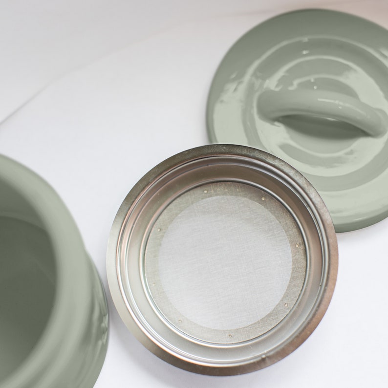 May include: A silver coffee filter with a mesh insert sits on a white surface. A sage green ceramic lid with a handle is also visible.