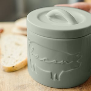 May include: A ceramic container with a lid and a pig design. The container is light green and has the word "bacon" embossed on the side.