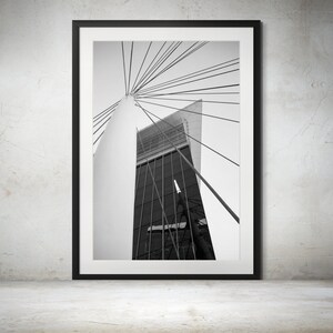 May include: Black and white photograph of a modern building with a cable-stayed bridge structure. The building is tall and slender, with a glass facade. The cables of the bridge are visible in the foreground, and the sky is visible in the background.