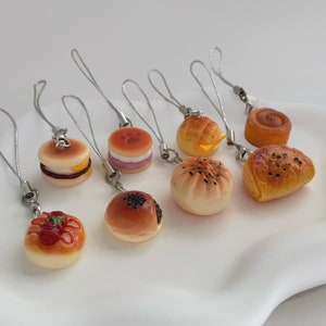 May include: A set of eight miniature food charms, each featuring a different type of bread or pastry. The charms are made of a light brown, beige, and white material and have a realistic appearance. They are attached to silver chains and would make a cute addition to a keychain or bag.