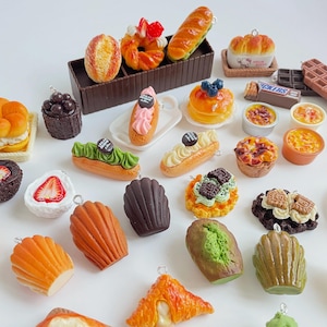 Peut inclure: Assortiment de charmes miniatures en pâte polymère représentant des aliments, notamment des pâtisseries, des pains et des desserts. Les charmes sont peints dans différentes couleurs, notamment le brun, le vert, l'orange et le blanc. Les charmes sont présentés sur une surface blanche.