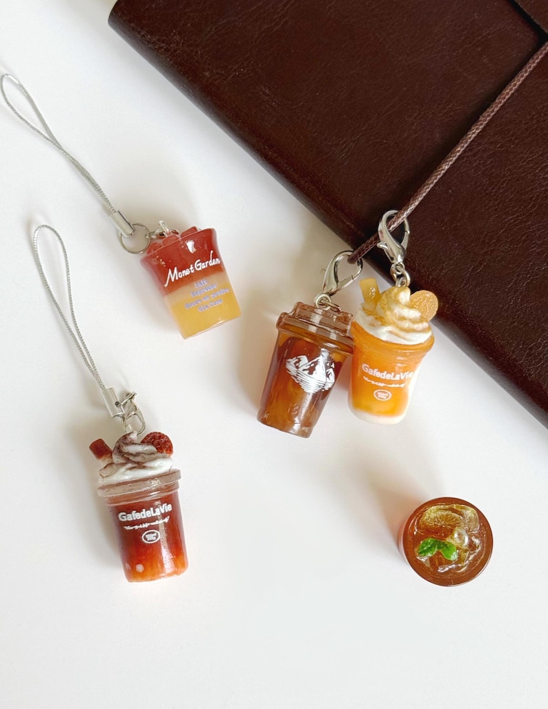 Puede incluir: Una colecci&oacute;n de dijes en miniatura con forma de bebidas heladas en vasos de pl&aacute;stico transparente. Los dijes incluyen etiquetas con los textos Monet Garden y GafedelaVie, con detalles realistas como crema batida, rodajas de fruta y menta.