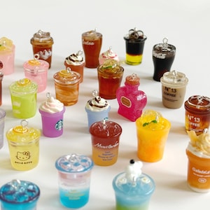Puede incluir: Una colecci&oacute;n de dijes en miniatura con forma de bebidas en vasos de pl&aacute;stico transparente, incluyendo caf&eacute;s helados, t&eacute;s frutales y refrescos en capas. Cada dije tiene una peque&ntilde;a argolla met&aacute;lica en la parte superior, con algunos vasos con texto.