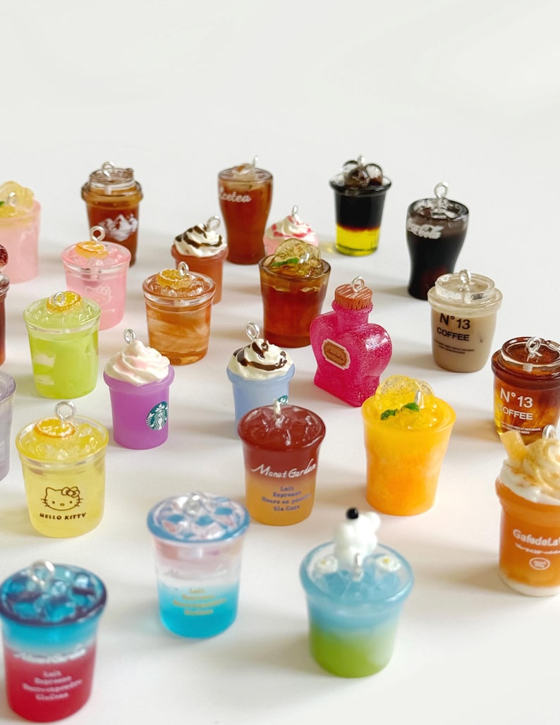 Puede incluir: Una colecci&oacute;n de dijes en miniatura con forma de bebidas en vasos de pl&aacute;stico transparente, incluyendo caf&eacute;s helados, t&eacute;s frutales y refrescos en capas. Cada dije tiene una peque&ntilde;a argolla met&aacute;lica en la parte superior, con algunos vasos con texto.