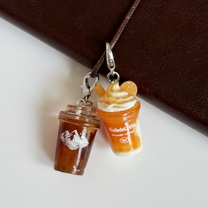 Puede incluir: Dos dijes de bebidas en miniatura con cierres de langosta plateados. Un dije es una taza de caf&eacute; helado marr&oacute;n oscuro con un gr&aacute;fico de monta&ntilde;a blanco. El otro es una bebida naranja con crema batida blanca, una rodaja de naranja y el texto GafedeLaVie.