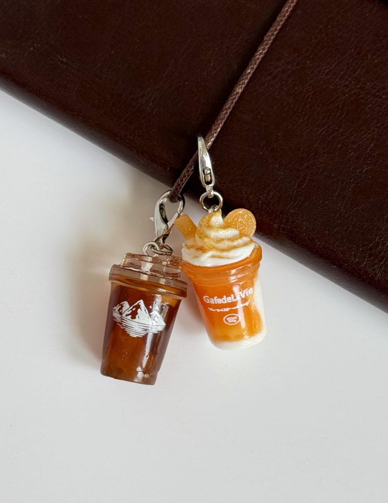 Puede incluir: Dos dijes de bebidas en miniatura con cierres de langosta plateados. Un dije es una taza de caf&eacute; helado marr&oacute;n oscuro con un gr&aacute;fico de monta&ntilde;a blanco. El otro es una bebida naranja con crema batida blanca, una rodaja de naranja y el texto GafedeLaVie.
