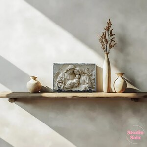 May include: A white stone relief sculpture of the Holy Family, depicting Mary, Joseph, and baby Jesus. The sculpture is mounted on a black slate base and displayed on a wooden shelf with two ceramic vases.