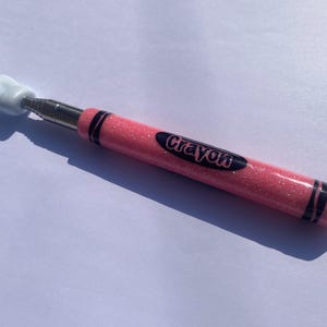 May include: A pink glittery crayon-shaped tool with the word "Crayon" on the side. The tool has a white hand-shaped end and black stripes. The tool is designed to be a novelty item.