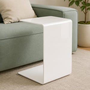 Könnte beinhalten: Ein weißer C-förmiger Beistelltisch mit einer glatten, glänzenden Oberfläche. Der Tisch hat ein durchgehendes, geschwungenes Design, das eine flache Tischplatte und einen vertikalen Stützfuß bildet, um ihn über ein Sofa zu schieben.