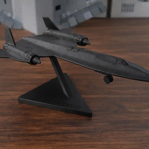Puede incluir: Un modelo de avión negro SR-71 Blackbird sobre un soporte negro. El modelo tiene detalles, incluyendo motores y alas. El soporte es triangular y tiene texto. El modelo se exhibe sobre una superficie de madera.