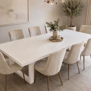 May include: A rectangular white dining table with a glossy finish and a sturdy base, surrounded by eight upholstered chairs in a light beige fabric. The chairs feature slim, metallic gold-toned legs and vertical stitching on the backrests.