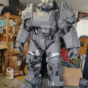 Puede incluir: Un traje de armadura asistida gris y negro de cuerpo completo, probablemente para cosplay o exhibición. El traje presenta un casco con respirador, hombreras y extremidades articuladas. La armadura está detallada con paneles y remaches, en un garaje.