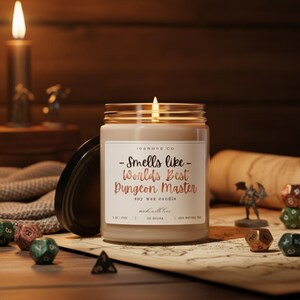 Puede incluir: Una vela de cera de soja encendida en un tarro de cristal con tapa negra. La etiqueta dice "Smells like Worlds Best Dungeon Master". Dados y figuras en miniatura están esparcidos alrededor de la vela sobre un mapa.