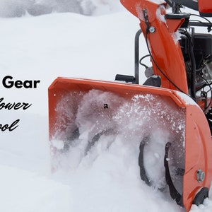 Op de afbeelding: Een rode sneeuwschuiver met een zwarte motor en wielen ruimt sneeuw van een oprit. De sneeuwschuiver werpt sneeuw uit de uitwerpkanaal. De tekst "Garage Gear Snowblower School" is zichtbaar in de afbeelding.