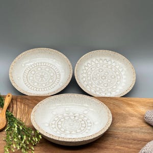 Könnte beinhalten: Drei weiße Keramikschalen mit geprägten Designs. Die Schalen stehen auf einer Holzoberfläche mit einem kleinen Holzlöffel und einer grünen Pflanze.