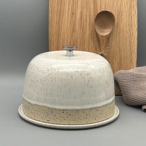 Könnte beinhalten: Eine Käseglocke aus Keramik mit gesprenkelter weißer und hellbrauner Glasur. Die Kuppel hat einen silbernen Griff und steht auf einem passenden Teller. Ein Holzschneidebrett und ein Löffel befinden sich im Hintergrund.